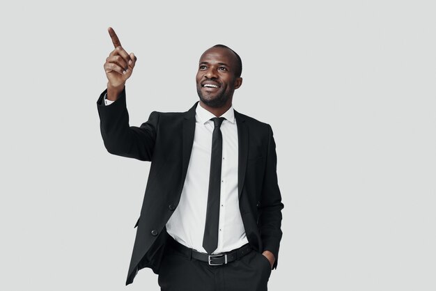 Handsome young African man in formalwear pointing copy space and smiling while standing against grey wall