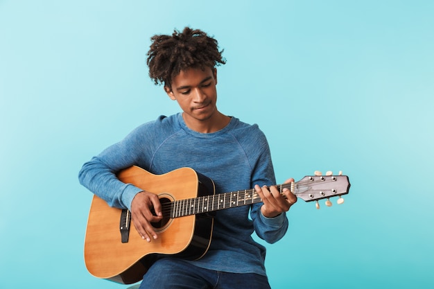 Handsome young african man playing a guitar while sitting isolated over blue