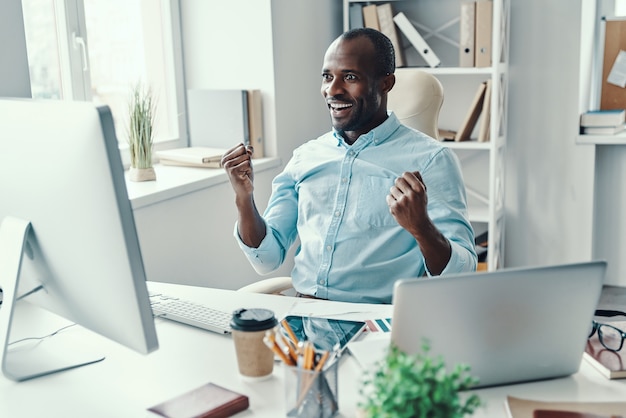 Bel giovane africano in camicia esulta e sorride mentre lavora in ufficio