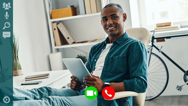 Handsome young african man in smart casual wear holding digital
tablet and smiling while having video call from home