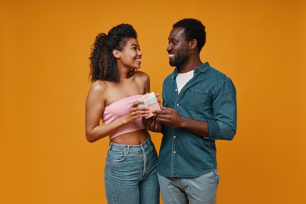 Handsome young African men giving a gift box to his girlfriend while standing against yellow background