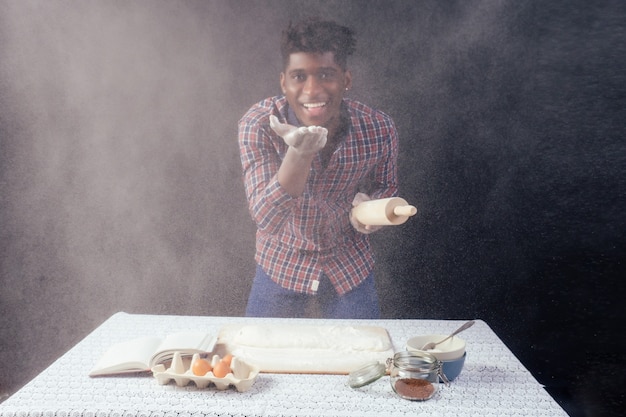 Handsome and young afro african man preparing homemade cakes American Pie from fresh dough hands dirty by flour, on the table are eggs, rolling pin and recipe book on a black background in the studio