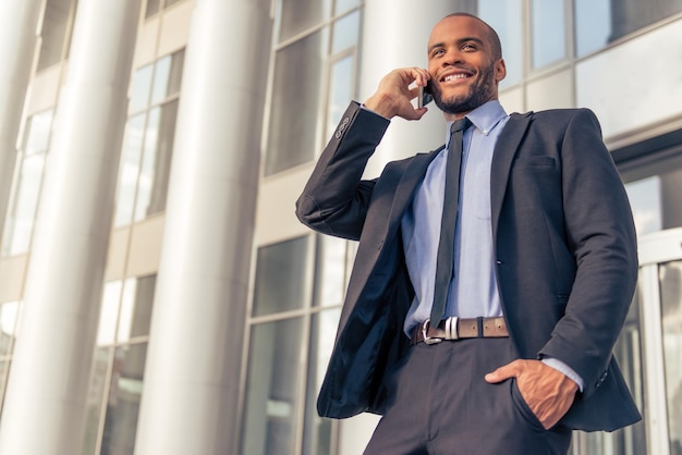 Handsome young Afro American businessman in classic suit talking on the mobile phone and smiling standing outdoors near the office building