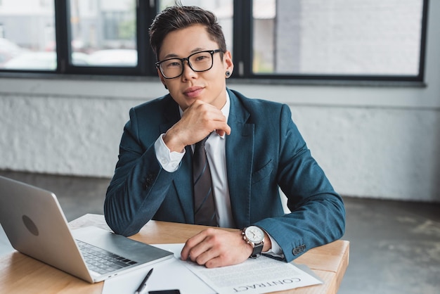 Handsome young asian businessman in suit and eyeglasses sitting at workplace and smiling at camera