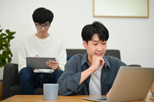A handsome young Asian male college student using a laptop in the living room with his friend