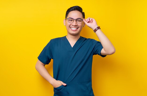 Handsome young asian man doctor or nurse wearing a blue uniform and glasses confident looking at the camera with smiling friendly isolated on yellow background healthcare medicine concept