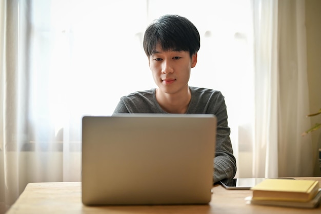 그의 집에서 노트북을 사용하여 노트북에 대한 학교 프로젝트에 집중하는 잘 생긴 젊은 아시아 남자