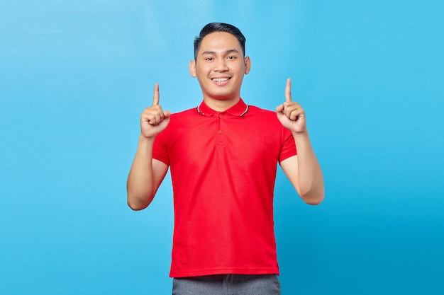 Handsome young Asian man in red shirt smiling pointing fingers up, inviting customers to special event isolated over blue background