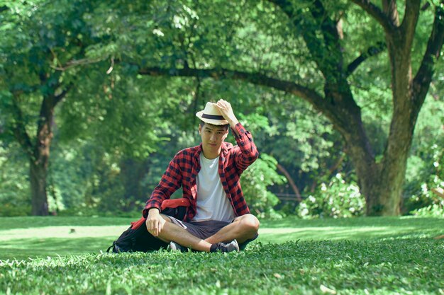 A handsome young Asian man sits on the lawn in the summer at the park.