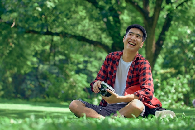 A handsome young Asian man sits on the lawn in the summer at the park.