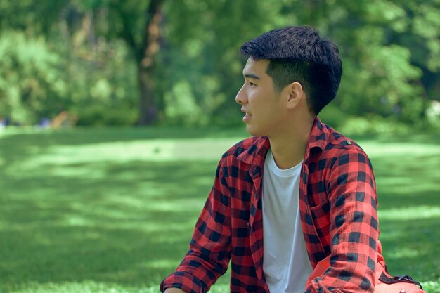 A handsome young Asian man sits on the lawn in the summer at the park.