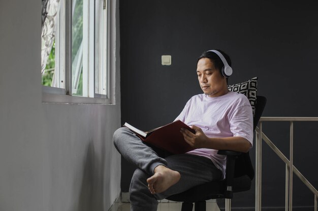 Bel giovane uomo asiatico seduto casualmente a leggere un libro mentre si gode la musica in appartamento