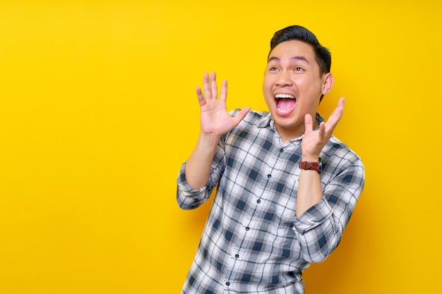 Handsome young Asian man surprised in plaid shirt looks aside and spreads his hands say wow isolated on yellow background people lifestyle concept