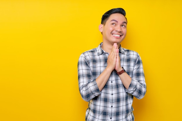 Handsome young Asian man trustfully pleaded in plaid shirt while hands folded in a prayer gesture begging about something isolated on yellow background people lifestyle concept