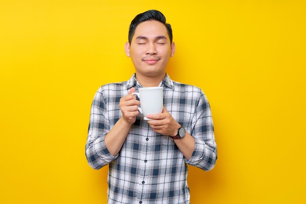 Handsome young asian man wearing a white checkered shirt enjoying a cup of coffee isolated on yellow background people lifestyle concept