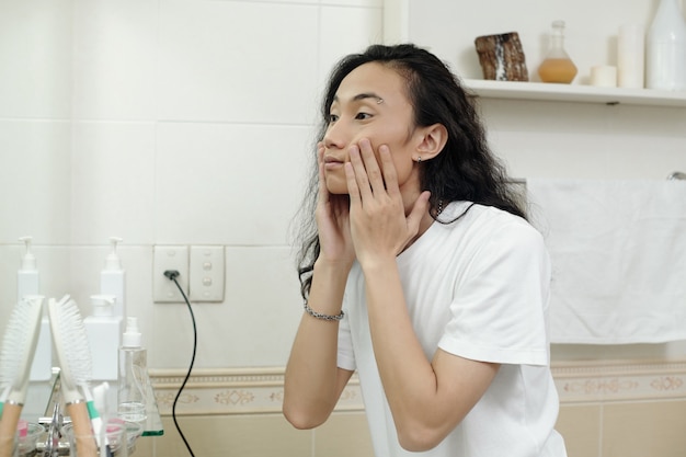 Handsome young Asian man with black wavy hair massaging cheeks while washing face in front of mirror in bathroom