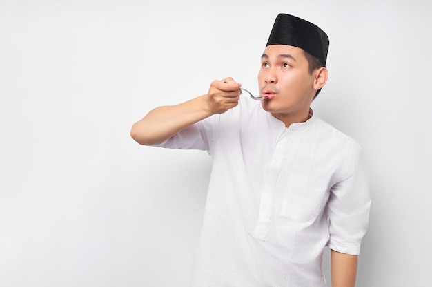 Handsome young Asian Muslim man holding spoon and tasting delicious food when breaking fast isolated on white background People religious Islamic lifestyle concept