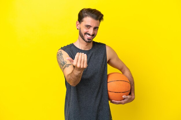 黄色の背景に孤立したハンサムな若いバスケットボール選手の男が手で来るように招待あなたが来たことを幸せ