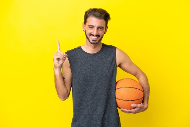 黄色の背景に分離されたハンサムな若いバスケットボール選手の男は、最高の兆候を示して指を持ち上げます