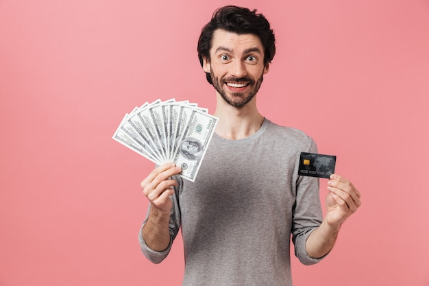 Handsome young bearded brunette man wearing sweater standing over pink, showing credit card, holding money banknotes