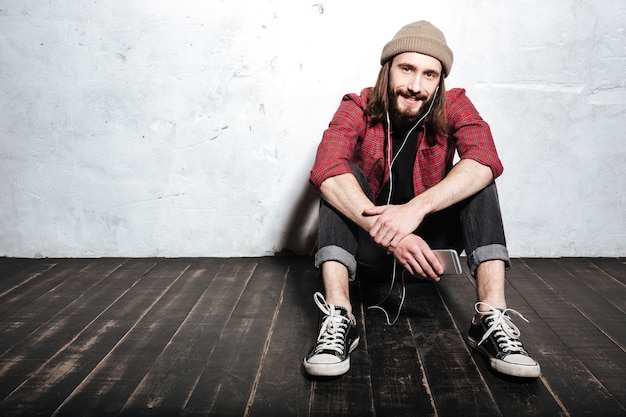 handsome young bearded hipster man wearing hat dressed in shirt in a cage sitting on floor isolated over wall while listening music with earphones