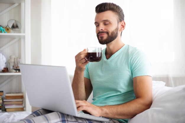 Красивый молодой бородатый мужчина в повседневной одежде работает дома, сидя на кровати. Уверенный в себе парень с ноутбуком и смартфоном пьет кофе в спальне.