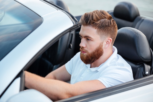 彼のカブリオレ車を運転しているハンサムな若いひげを生やした男