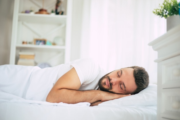 Handsome young bearded man is sleeping in a white big bed in a comfortable bedroom