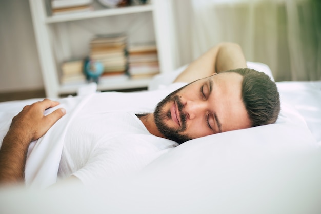 Handsome young bearded man is sleeping in a white big bed in a comfortable bedroom