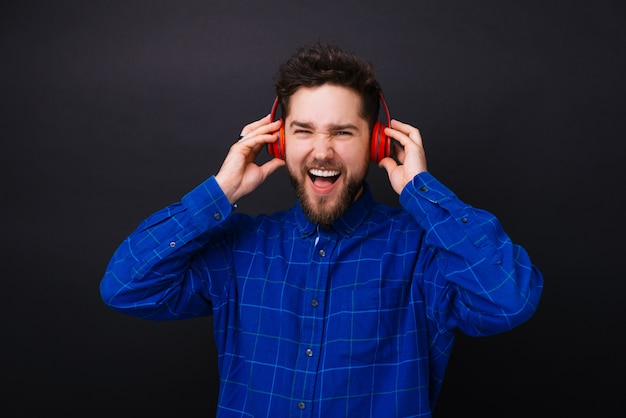 Handsome young bearded man listening to the music on headphones and singing on black background.