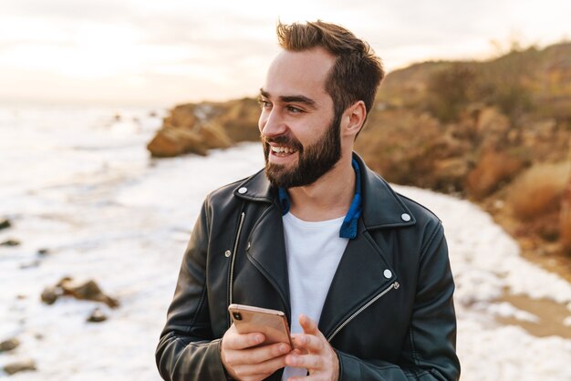 Handsome young bearded man wearing a jacket using mobile phone while standing at the beach