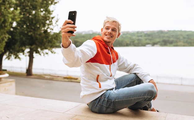 Handsome young blond man enjoying weather in city park waiting for his girlfriend texting message while listening to his favorite music from wireless headphone using smart phone outdoor in the park