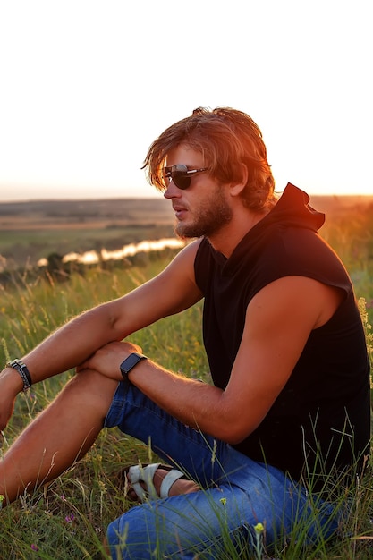 a handsome young blond man in glasses and a black Tshirt is sitting on the grass at sunset