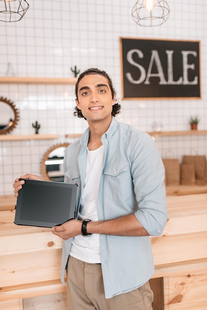 Handsome young boutique owner holding digital tablet with blank screen and smiling at camera