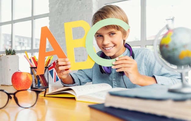 Handsome young boy holding colorful english letters while sitting in the light classroom