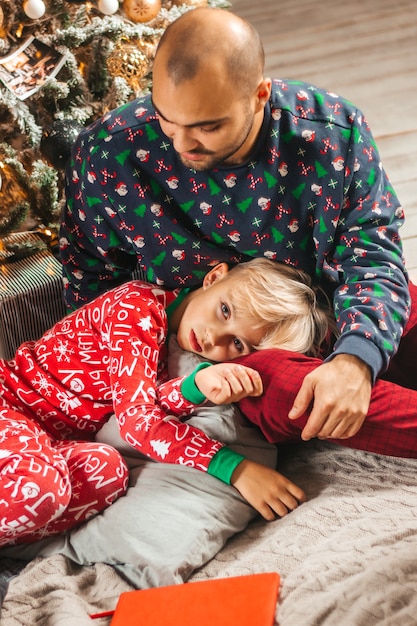 ハンサムな少年は、クリスマスのパジャマで彼のお父さんの膝の上に横たわっています。