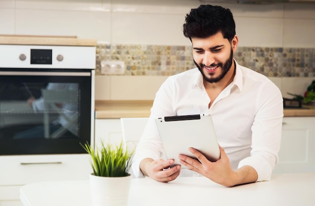 Handsome young boy with back wavy hair and neat beard using tablet in modern light kitchen wearing pristine white shirt