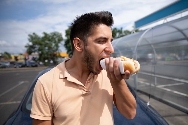 ガソリンスタンドの近くの駐車場でホットドッグを食べてハンサムな若いブルネットの男。