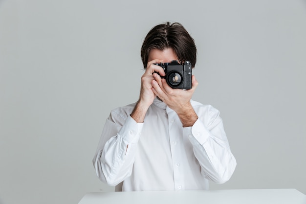 Handsome young brunette man sitting at the table and making with front isolated on the gray wall