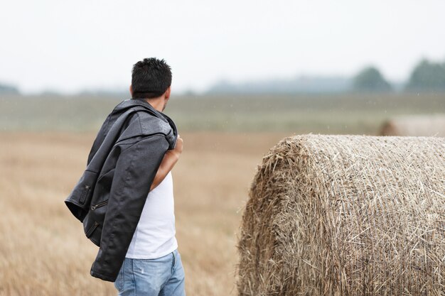 A handsome young brunette man stands in a field near a haystack.