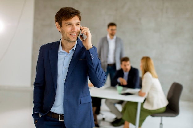 Handsome young business man standing confident in the office in front of his team and using mobile phone