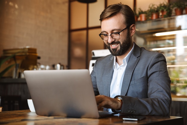 Handsome young businessman dressed in suit working on a laptop computer, sitting at the cafe indoors, holding mobile phone