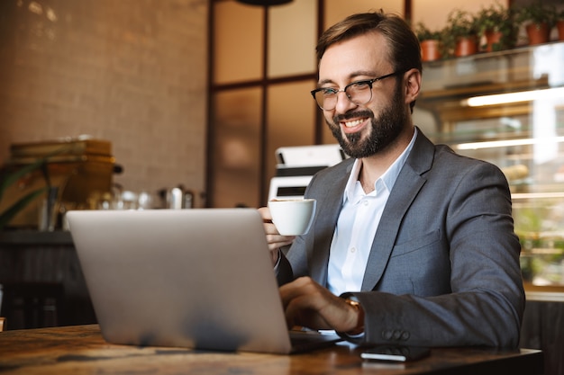 Handsome young businessman dressed in suit working on a laptop computer, sitting at the cafe indoors