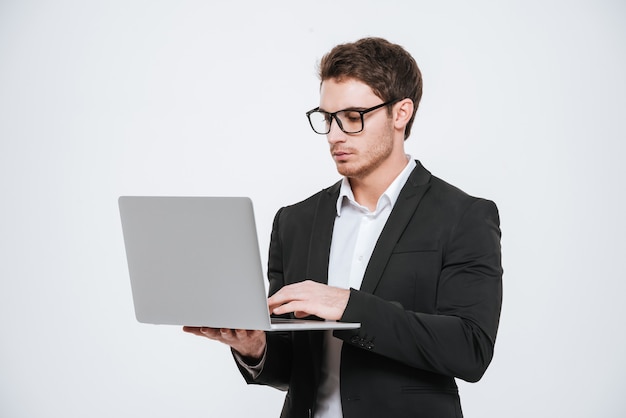 Handsome young businessman in eyeglasses standing and using laptop over white wall