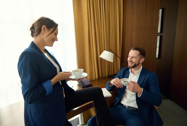 Handsome young businessman sitting on an armchair and laughing while talking to his handsome partner over a cup of coffee in a hotel room during a business trip. Business people on coffee break