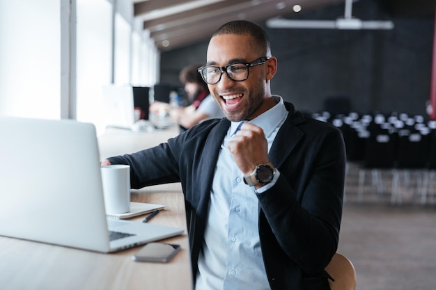 Bel giovane uomo d'affari seduto alla sua scrivania, celebrando il successo con le braccia alzate mentre guarda lo schermo del suo laptop