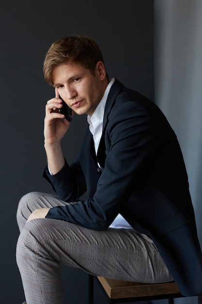 Handsome young businessman in suit sitting and looking at camera, holding his phone near his ear, over black background.