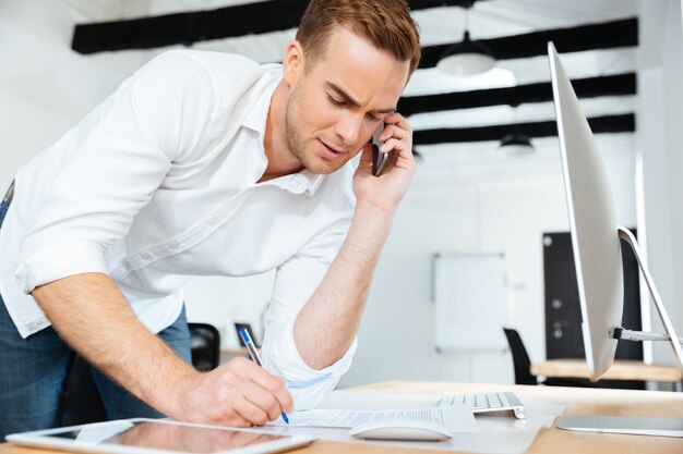 Handsome young businessman talking on cell phone and writing in office