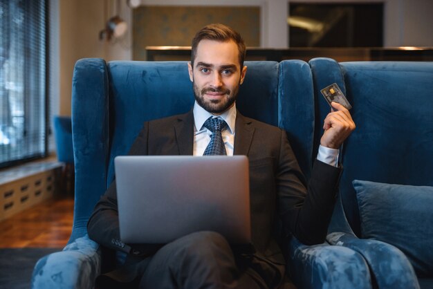 Handsome young businessman wearing suit sitting at the hotel lobby, using laptop computer, showing plastic credit card