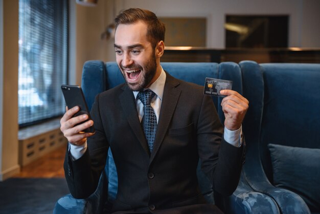 Handsome young businessman wearing suit sitting at the hotel lobby, using mobile phone, showing plastic credit card